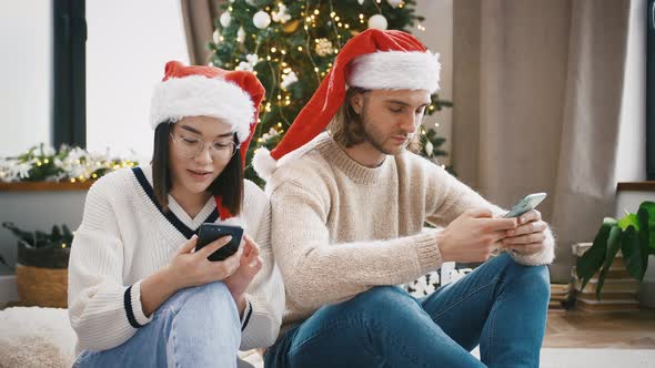 Multiracial Couple in Santa Hats are Laughing Using Cellphones Sitting on the Floor at Home Against