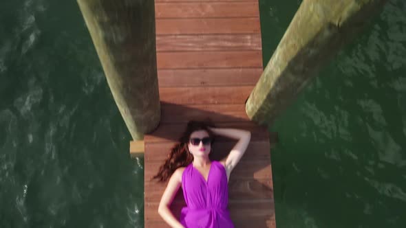 Aerial View of Sexy Girl With Long Legs in Purple Dress Lying on Wooden Dock Above Sea Drone Shot