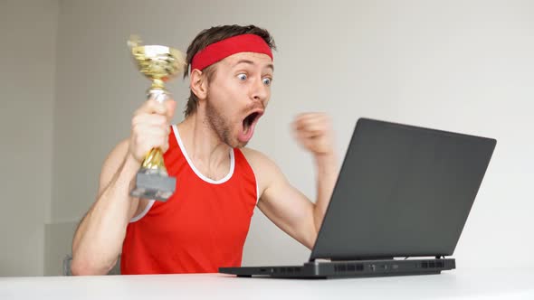 Sportsman celebrating victory with golden cup while watching result on laptop.