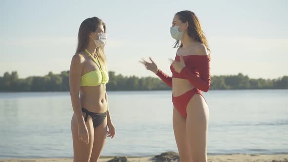 Two Gorgeous Tanned Slim Women in Face Masks and Swimsuits Talking on River Bank at Sunset. Side