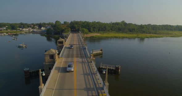 Lowering Aerial Pan of Cars Driving on Drawbridge Over Water and Near a Forest