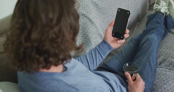 Caucasian man sitting on sofa in living room using smartphone and drinking coffee