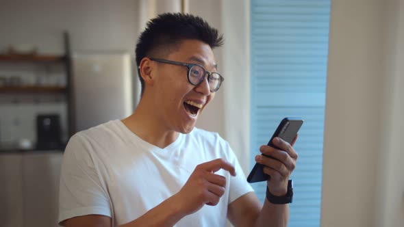 Handsome Asian Young Man Standing Near Window Reading Email on Mobile Phone Making Winner Gesture