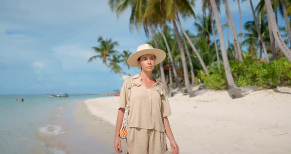 Front Shot of Brunette Woman  Dressed in Safari Look and a Hat Walks on Empty Tropical Beach
