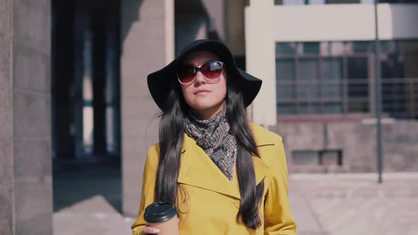 Girl in a Yellow Raincoat Hat and Glasses Walking Down the Street and Drinking Coffee