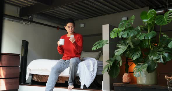 Man with coffee cup using mobile phone on bedroom
