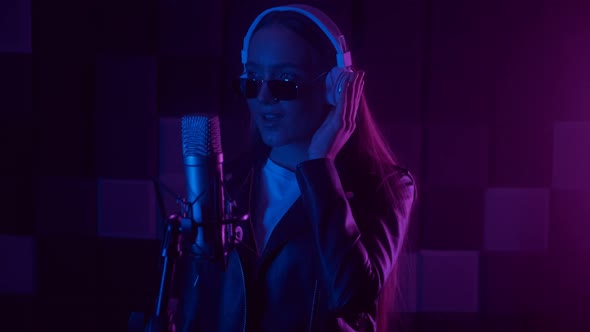 Attractive Woman in Glasses and Headphones Sings Into a Microphone in Neon Light in the Dark Studio