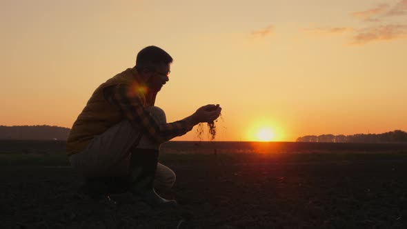 A Man Agronomist Examines the Soil in His Hands in a Field at Sunset