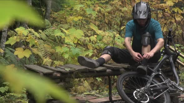 A young man mountain biker tying his shoes while resting by his bike in a forest.