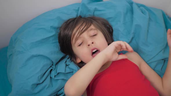 Sick Boy a 5Yearold Child with Long Black Hair Sneezes Into His Elbow While Lying on the Bed