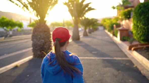 Woman Runs Down the Street Among the Palm Trees at Sunset Back View