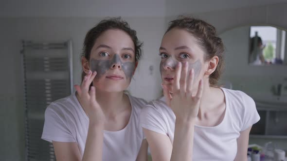 Slim Charming Caucasian Twin Sisters Touching Skin Adjusting Face Mask Looking at Camera As Mirror
