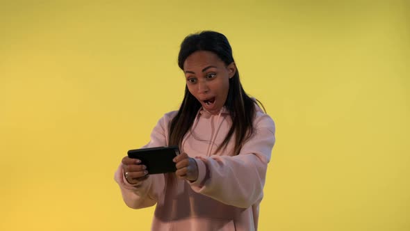 Pleasant-looking Black Girl Playing Games on Smartphone on Yellow Background.