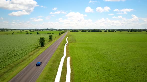 Aerial view following an electric blue car zooming down a gravel road in the countryside