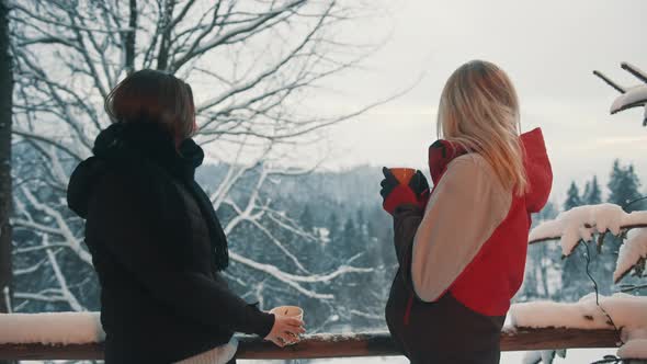 Two Young Women Enjoying View of the Mountain Covered in Snow and Drinking Hot Beverage on Winter