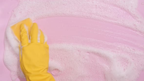 Hand in a Yellow Rubber Glove Holds a Cleaning Sponge and Wipes a Soapy Foam on a Pink Background