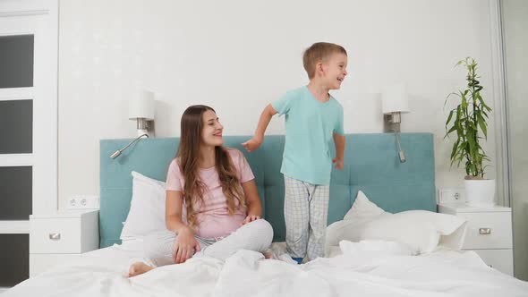 Beautiful Young Woman in Pajamas Looking on Her Little Toddler Son Jumping on Her Bed at Morning