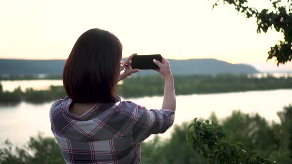 A Young Woman Stands on a Hill Against the Background of the River and Mountains and Photographs the