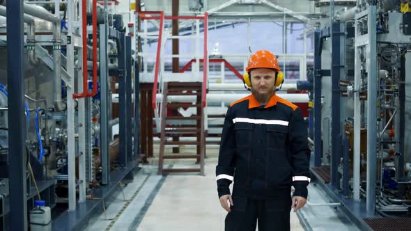 A Plant Worker in a Hard Hat and Earphones Stands in a Gas Compression Shop Near a Working