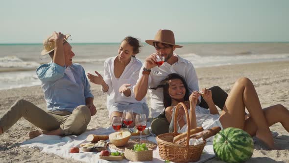 Four Happy Millennial Friends Have Picnic on Sandy Ocean Beach at Sunny Day Next Waving Sea