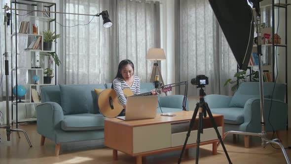 Asian Woman Reading Comments On Laptop And Speaking To Camera While Live Stream Playing A Guitar