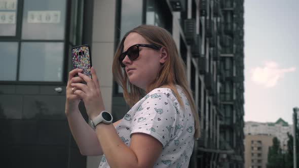 Young Married Girl in Glasses Makes a Photo Video on Phone Against the Background of New High House