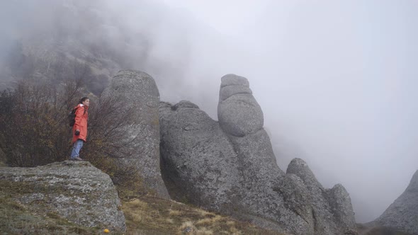 Woman Stands on Steep Cliff Looking at Rocks Against Mist