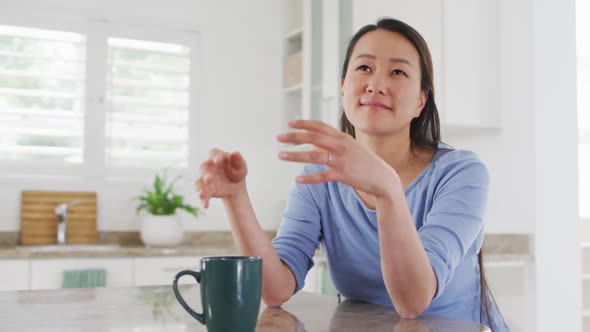 Happy asian woman sitting in kitchen and drinking coffee