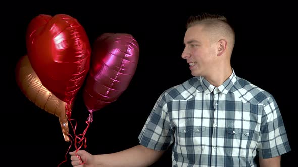 A Young Man Is Standing with Heart-shaped Balloons. Man Holds Helium Balls in His Hands on a Black