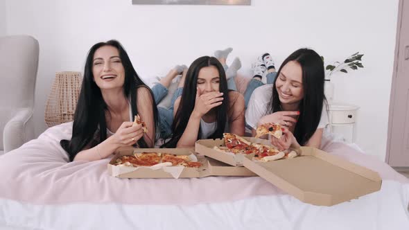 Three Gorgeous Girlfriends Are Lying on the Bed and Eating Delivery Pizza