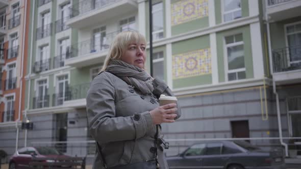 Confident Senior Caucasian Woman Walking Along City Street and Drinking Coffee. Relaxed Female