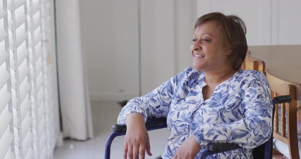 Happy african american senior woman in wheelchair looking out of window at home smiling