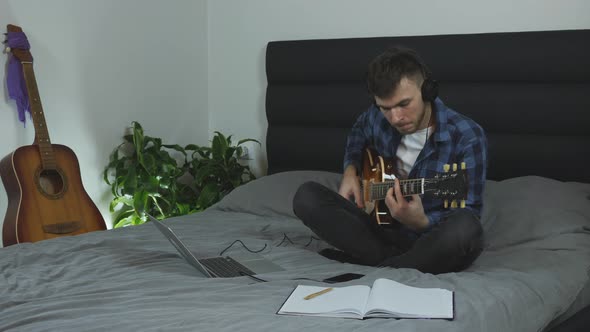 Musician in headphones playing on electric guitar on bed in living room. Music concept