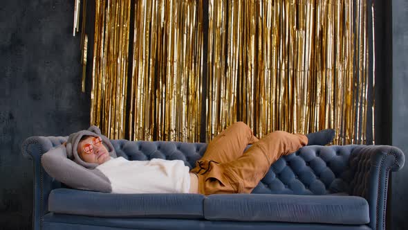 A Young Man in a Hood with Ears and Bright Glasses is Lying on a Sofa Against a Wall with Golden