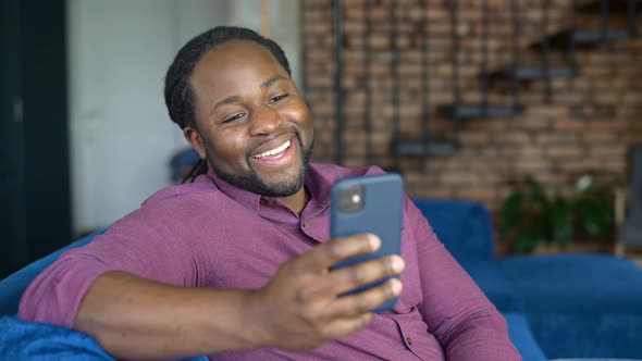 Smiling AfricanAmerican Man Using Smartphone Sitting on the Sofa at Home