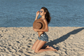 Dark-haired girl in jean shorts kneels in sunglasses on the sand near the sea and hold a hat on her - PhotoDune Item for Sale