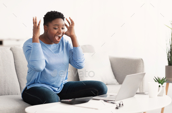 rvy woman happy with money win, satisfied about success. Black lady screaming yes and looking at laptop computer, sitting on the couch at home