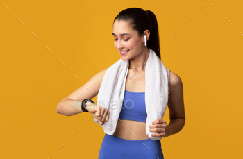 With Towel On Neck Looking At Wearable Bracelet, Checking Activity Tracker, Wearing Wireless Earbuds Isolated On Orange Studio Background
