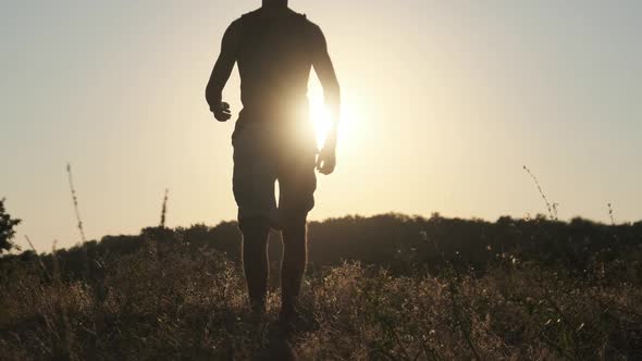 Silhouette of Young Man Walking in Field To Setting Sun and Raising Hands Up