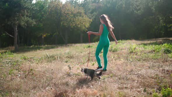 Woman in a sporty turquoise overalls walks with her fluffy cat on a leash in the forest