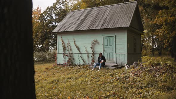 Woman Is Sitting on the Porch Near Small House Outside the City, Reading a Book