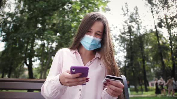 Girl in Protective Medical Mask with Credit Card and Phone Makes Online Payments