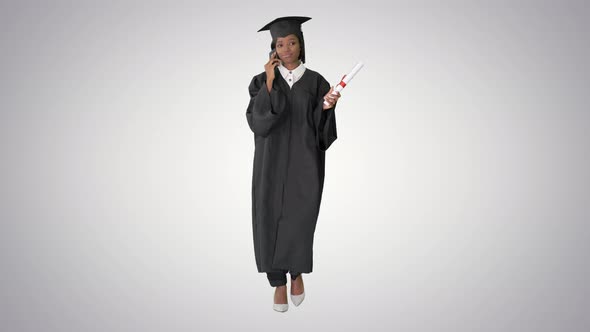 African American Female Student in Graduation Robe Talking on the Phone While Walking on Gradient