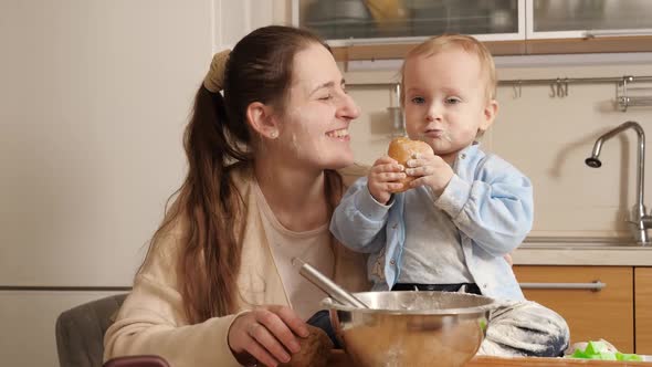 Happy Smiling Mother Hugging Her Little Baby Son Sitting on Kitchen Table While She is Baking Home