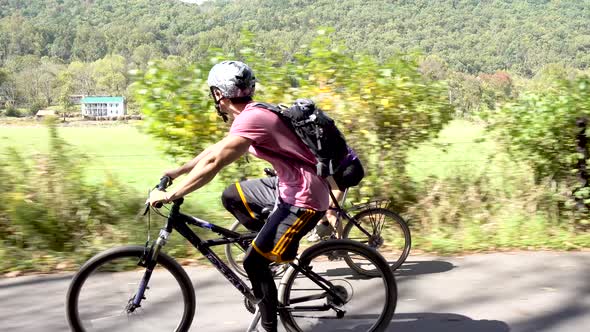 Mother and teenage son biking side by side on a rural road with a farm in the background.