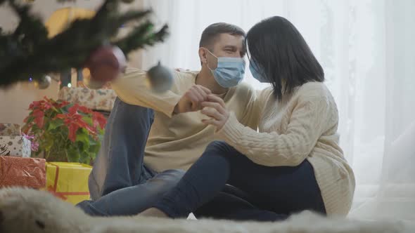 Relaxed Caucasian Couple in Covid19 Face Masks Sitting at New Year Tree at Home and Hugging