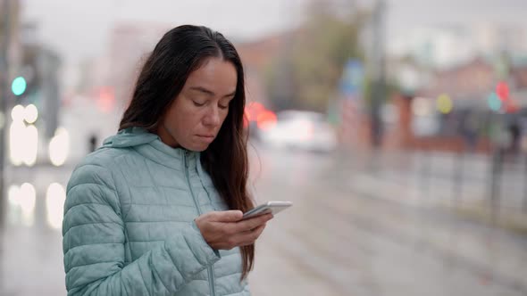 a Brunette in a Blue Jacket and with Long Hair Stands with a Phone in Hand and Looks at the Screen