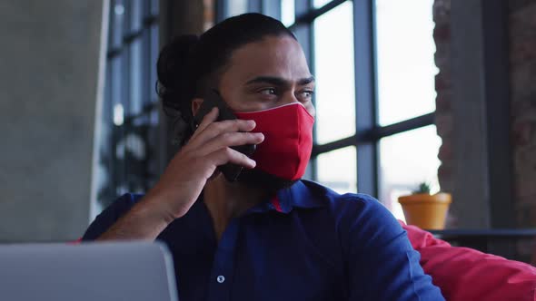 African american man wearing face mask sitting in cafe talking on smartphone using laptop