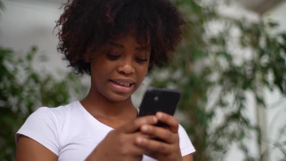 Closeup Lowangle View of Cheerful AfricanAmerican Young Woman Holding Smartphone in Hands Enjoying