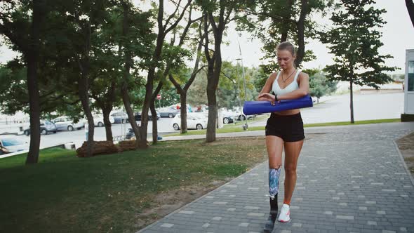 Young Girl with Leg Prosthesis Going to Training with Fitness Mat Looking at Smartwatch